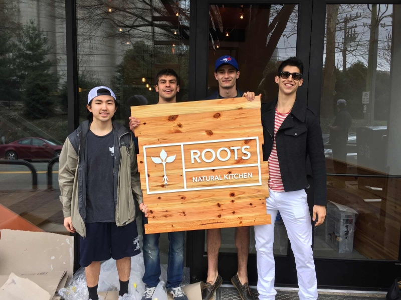 Four people standing outside a storefront holding a wooden “Roots Natural Kitchen” sign
