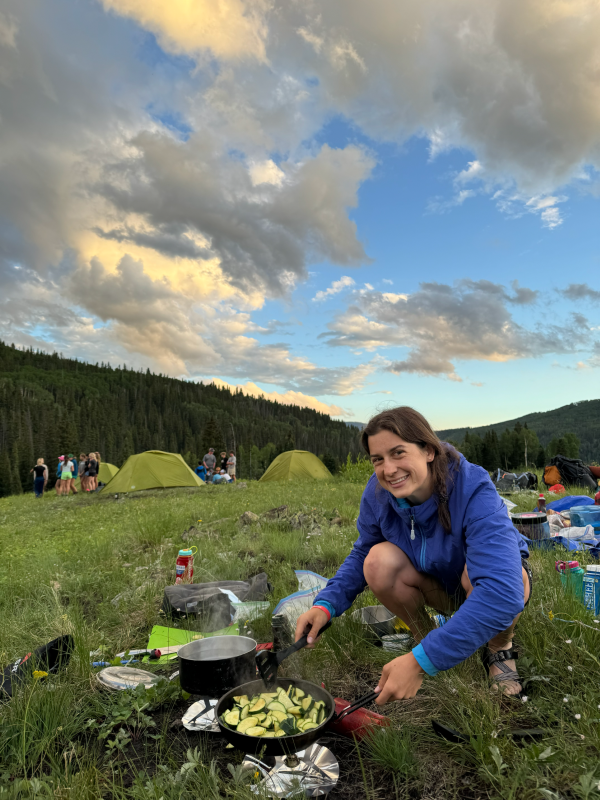 Emily Bronstein cooking at a campfire, with fellow campers, tents, and mountains in the background