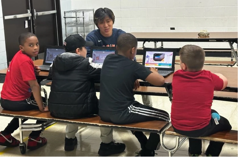 David Sun sitting at a school cafeteria table, talking to four elementary school students